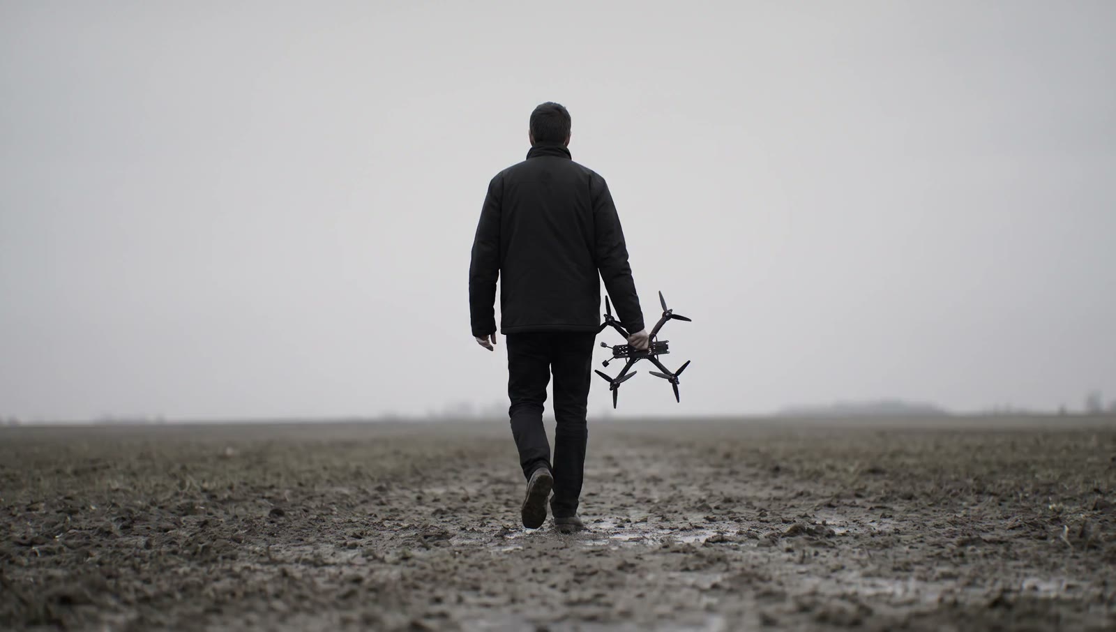 One person walking away across a bare ploughed field under flat overcast, carrying a small exposed-frame quadcopter at their side